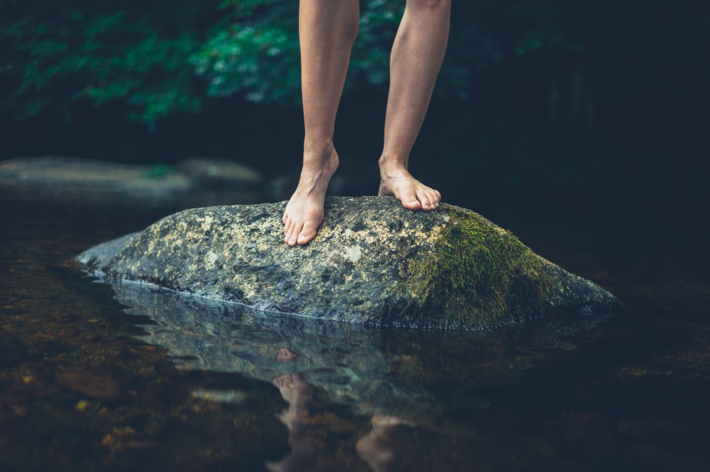 A person standing barefoot on a moss-covered rock in a river, surrounded by greenery.