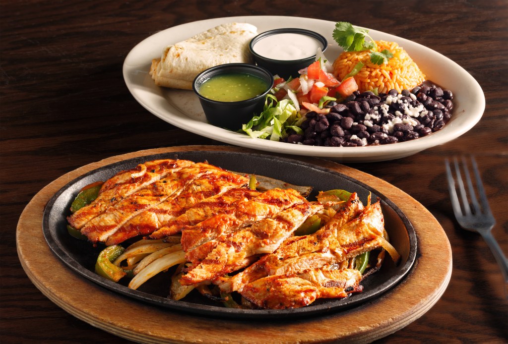 Sizzling grilled chicken served with sautéed peppers and onions on a hot plate, accompanied by a side of rice, black beans, lettuce, salsa, and tortillas.