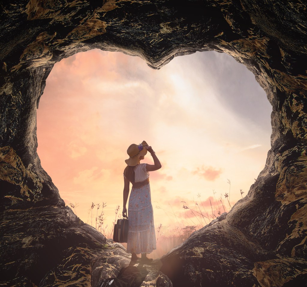 A silhouette of a person wearing a hat and holding a suitcase stands in a cave's heart-shaped opening, gazing at a colorful sunset sky.