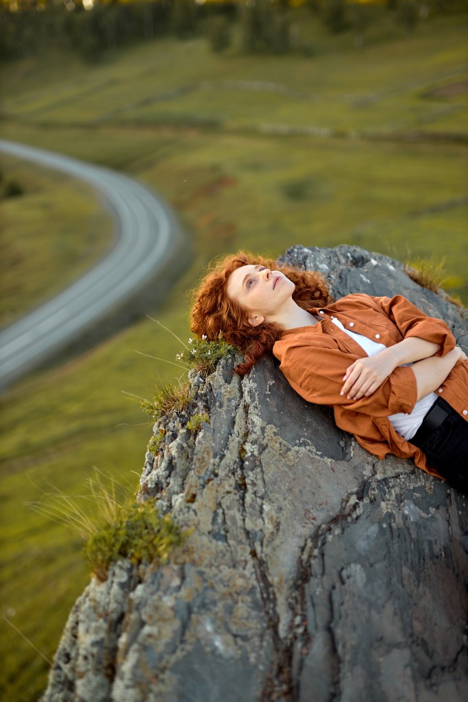 A woman with curly red hair lying on a rock, looking up at the sky, with a winding road and green fields in the background.
