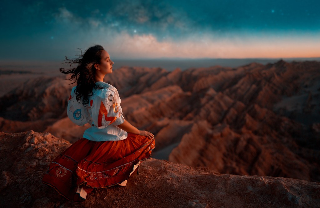 A woman sitting on the edge of a rocky cliff, wearing a colorful traditional dress, gazes thoughtfully at the sunset over a mountainous landscape.