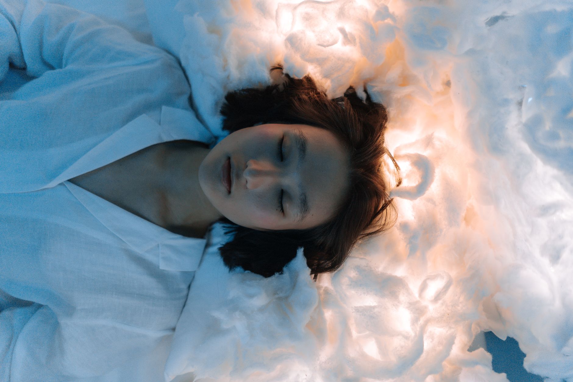 A serene woman with closed eyes resting on a soft, cloud-like surface illuminated by gentle light.
