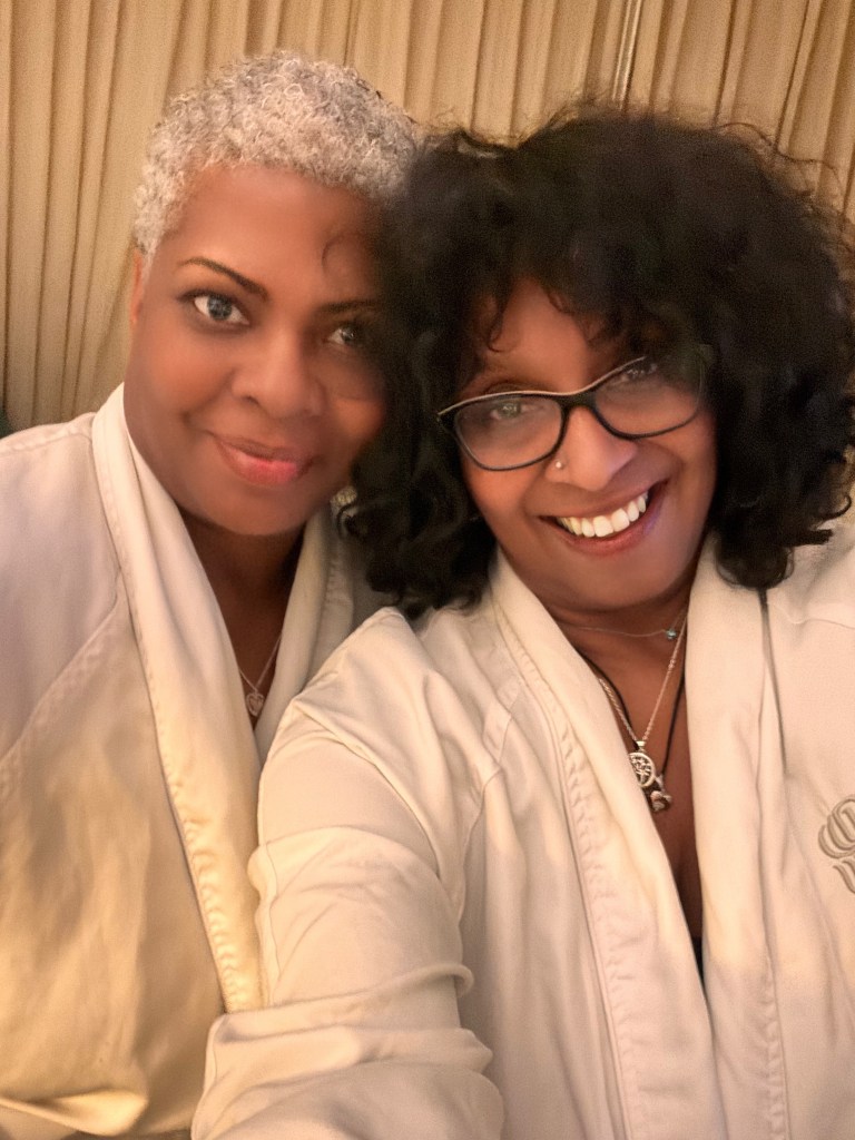 Two women posing together, wearing beige robes, smiling at the camera.