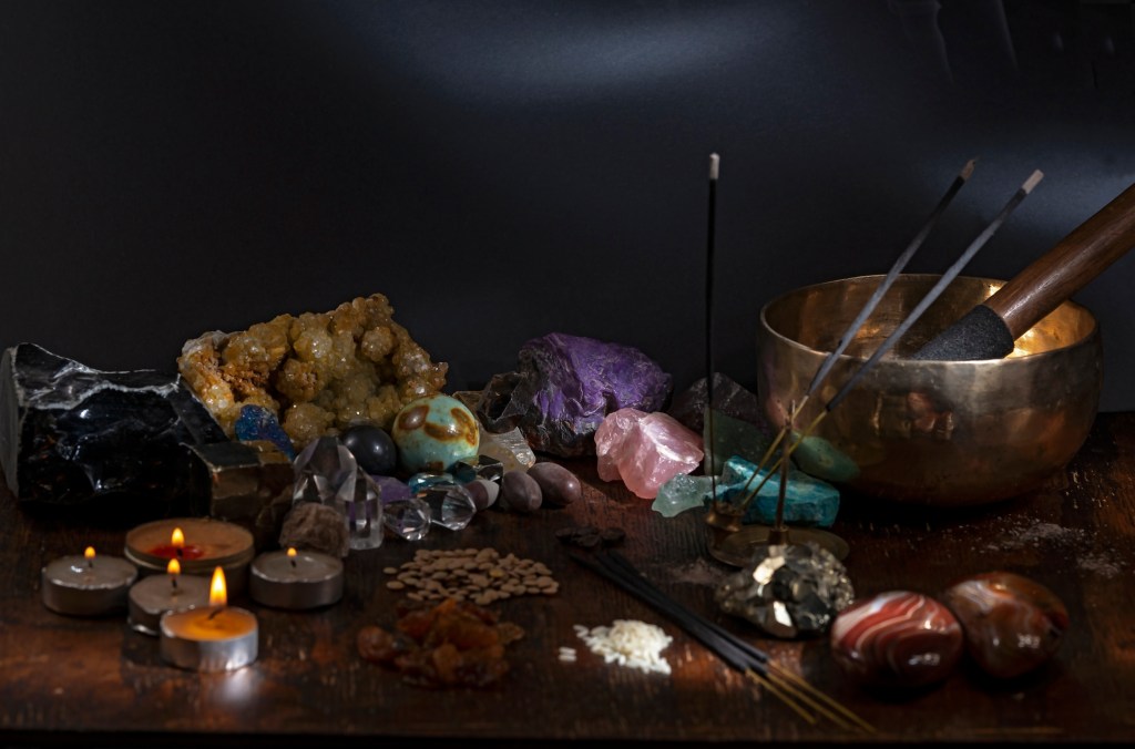 A collection of crystals, gemstones, and minerals arranged on a wooden surface, accompanied by candles, incense sticks, and a singing bowl.