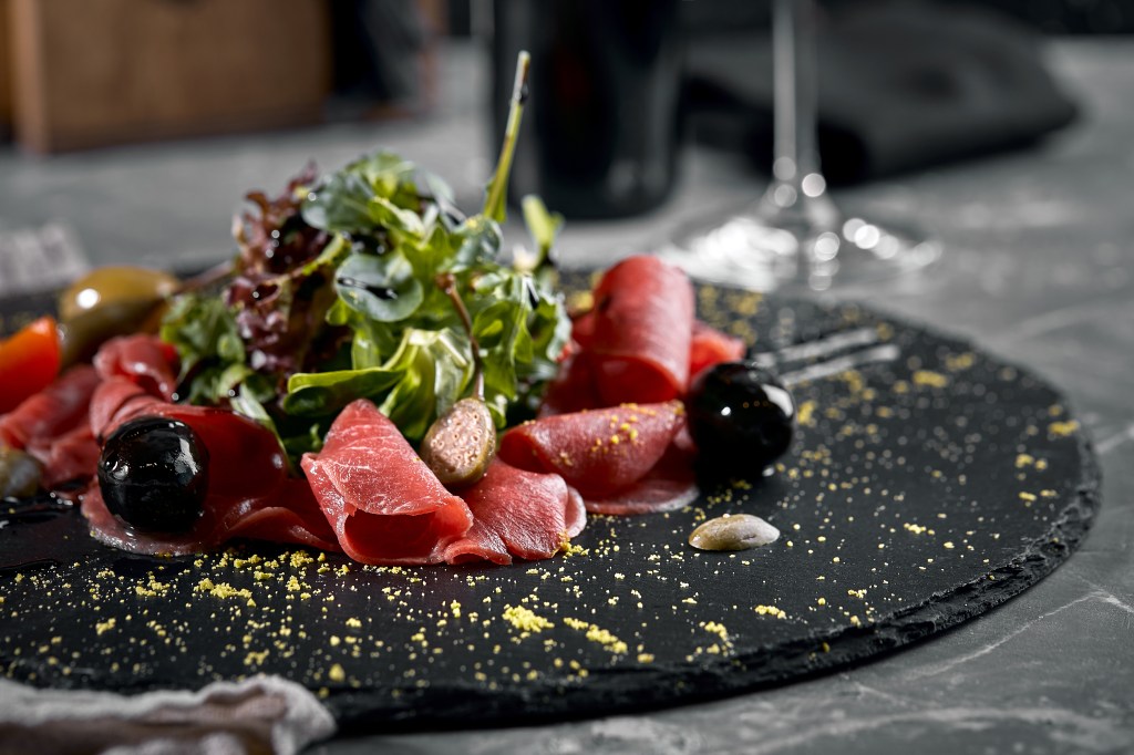 A gourmet platter featuring thinly sliced cured meat, black olives, green salad leaves, and sprinkled with yellow breadcrumbs on a black slate plate.