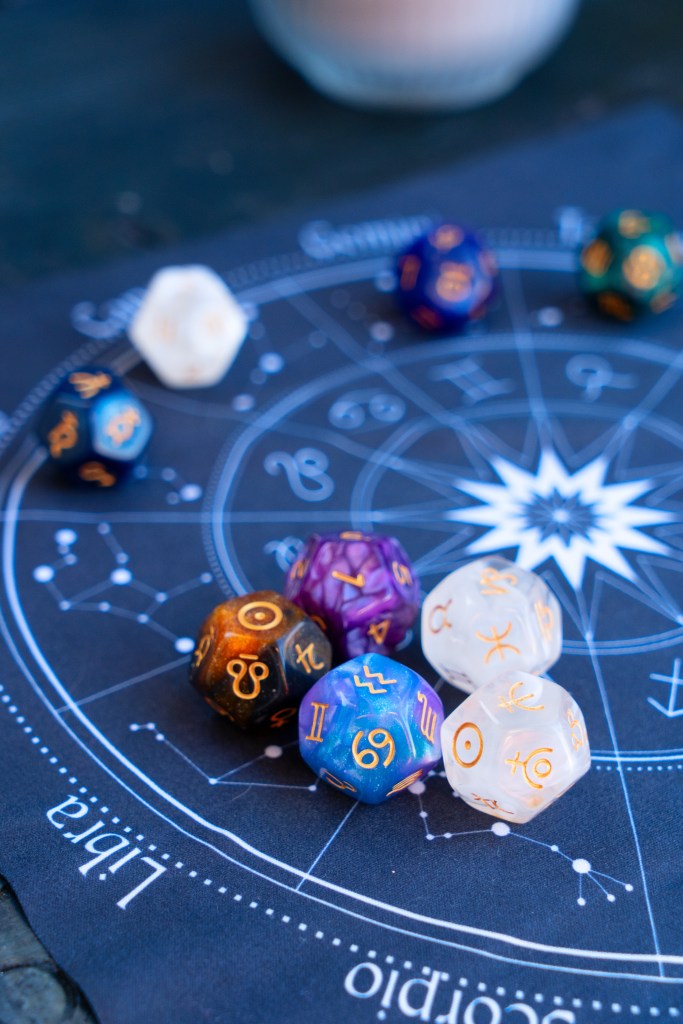 A collection of colorful astrological-themed dice arranged on a printed astrology chart, with symbols and zodiac signs visible.