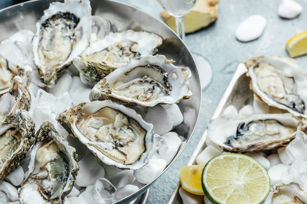 A bowl of fresh oysters served on ice, accompanied by lemon slices and a glass of sparkling drink.