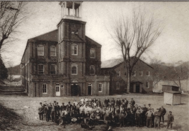 Historic black and white photo of a large group of people gathered in front of a multi-story building with a clock tower and a rural landscape in the background.