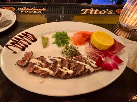 A plate of sliced seared tuna accompanied by wasabi, pickled ginger, and various vegetable garnishes including greens and carrot, drizzled with a creamy sauce and decorative sauce patterns.