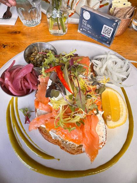 A plate featuring a slice of bread topped with cream cheese, smoked salmon, mixed greens, and slices of onion. Accompanied by pickled red onions, capers, a wedge of lemon, and a green sauce, with a small bill holder in the background.