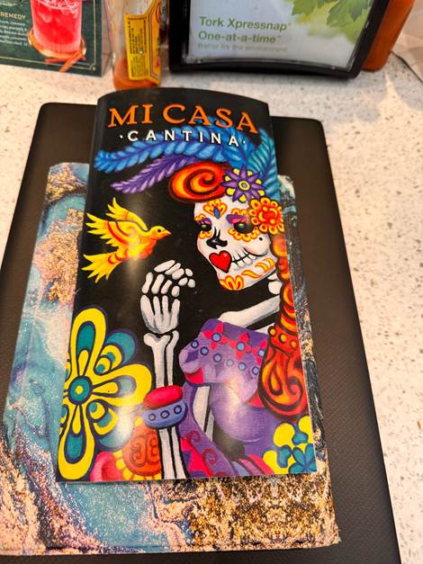 Colorful menu cover for Mi Casa Cantina featuring a decorated skull and vibrant flowers, placed on a table with a textured background.