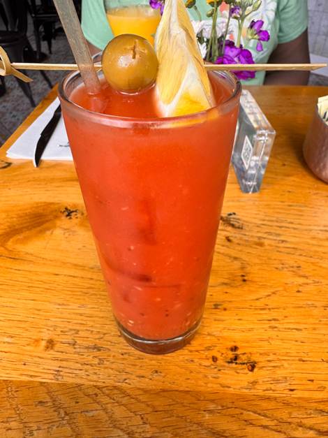 A tall glass of vibrant red cocktail garnished with an olive and a slice of lemon, resting on a wooden table.