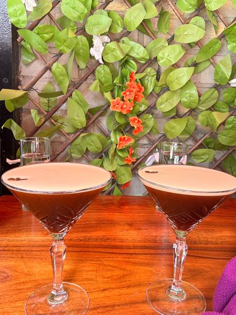Two elegant cocktails in martini glasses on a wooden table, surrounded by green leaves and orange flowers in the background.